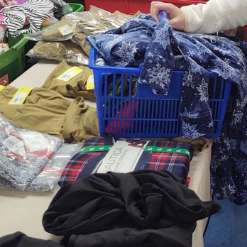 Cellar Dwellas - The image shows a table covered with different clothing items and accessories, including scarves, pants, and packaged garments. A blue plastic basket contains more scarves, and a plaid-patterned item is visible. Several people are seen browsing or shopping, with some holding or reaching for items. The setting appears to be a thrift store, clothing sale, or market. - Shoe Stores Near Me - Hampstead, New Hampshire