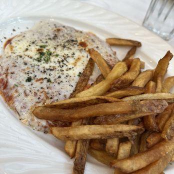 Gino's Restaurant - Chicken cutlet parm with fries and fresh pepper - A serving of chicken parmesan with melted cheese, herbs, and seasoning is presented on a white plate. The dish is accompanied by a generous portion of French fries, which are golden-brown and crispy. The background shows a table setting with cups, glasses, and utensils, indicating a restaurant dining environment. - Pizza Near Me - Brooklyn, New York