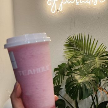 Teaholic - A person is holding a tall, clear cup filled with a layered pink and white beverage, topped with a white lid. The background features a wall with a neon sign that reads 'take a sip of paradise' and a large potted plant with broad, green leaves. The setting appears to be inside a cafe or similar establishment. - Bubble Tea Near Me - Cambridge, Massachusetts