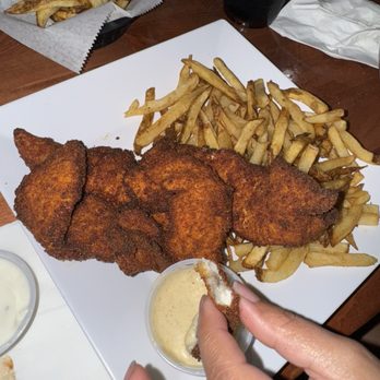 Straphanger Bar and Grill - A plate with fried fish fillets, French fries, and a small cup of dipping sauce is on a restaurant table, with a drink and additional fries in the background. - American Near Me - Hackensack, New Jersey