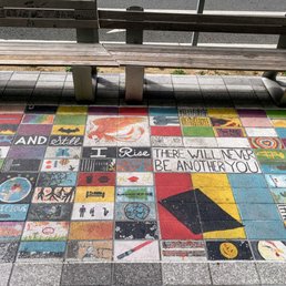 A section of sidewalk features a grid of colorful tiles with various artistic designs, patterns, and messages. Some tiles display images such as a fish, a globe, and abstract art, while others contain text including "AND still I Rise" and "There will never be another you." The tiles are positioned near a wooden bench with metal supports, and the area appears to be outdoors, possibly in an urban or park setting.
