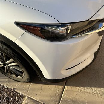 Bumper Buddies - After - A photograph captures the front left side of a white car, focusing on the headlight, part of the hood, and the front wheel. The car is parked on a concrete surface with a section of gravel visible nearby. The headlight has a sleek, modern design, and the tire features a detailed rim. The image is taken in natural daylight, highlighting the car's clean exterior and the surrounding ground. - Mobile Dent Repair Near Me - Phoenix, Arizona