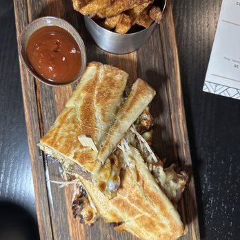 Rum Room - Pan con Bistec - A toasted sandwich with golden-brown bread, filled with melted cheese and meat, is presented on a wooden tray. It is accompanied by a small metal cup of French fries and a cup of dipping sauce, likely ketchup or barbecue sauce. The tray is placed on a dark surface, with part of a menu visible nearby. - Cocktail Bars Near Me - Miami Beach, Florida