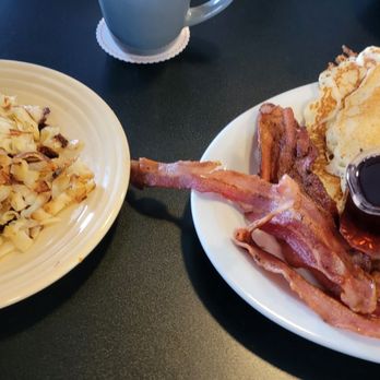 J’s Hideaway - A breakfast meal includes a stack of pancakes with butter, crispy bacon strips, and two small containers of syrup. The food is served on a white plate, placed on a dark table. The setting suggests a casual dining environment, likely a diner or breakfast restaurant. - Diners Near Me - Tehachapi, California