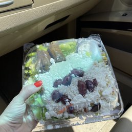 Mediterranean Salad - A person with red nail polish holds a plastic container of Mediterranean salad, including grape leaves and cheese, inside a vehicle.