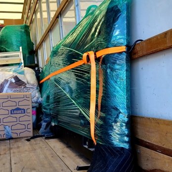 DV Movers - Tv beyond secure and locked in place - A large item, possibly a piece of furniture or equipment, is wrapped in green plastic and secured with orange straps inside a truck or storage area. The space has wooden flooring and walls with exposed framing, and other items such as boxes and bags are visible nearby. - Movers Near Me