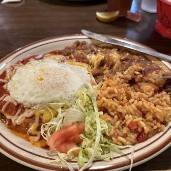 El Bruno's - Enchiladas!! - A ceramic plate holds a serving of Mexican food, including rice with tomato and spices, refried beans, and a fried egg placed on top of a layered dish with cheese and sauce. Garnished with shredded lettuce and tomato slices, the plate is set on a wooden table with utensils and condiments visible in the background. - Mexican Near Me - Albuquerque, New Mexico