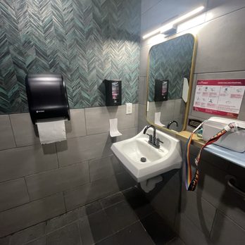 Command Coffee - Bathroom - The image shows a small bathroom with a wall-mounted white sink and a black faucet. Above the sink is a rectangular mirror with a gold frame, illuminated by a horizontal light fixture. The wall behind the sink has a decorative tile pattern, while the lower part is covered with plain gray tiles. To the left of the sink, there is a black paper towel dispenser, and to the right, a small shelf with a white paper towel holder. An electrical outlet is visible on the wall, and a colorful lanyard hangs from the shelf. - Coffee & Tea Near Me - Indianapolis, Indiana