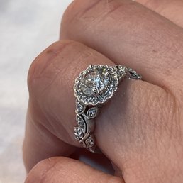A close-up image shows a diamond engagement ring with a large, round-cut center stone surrounded by a halo of smaller diamonds. The band has detailed metalwork with additional small diamonds embedded in a decorative pattern. The ring is worn on the person's finger, which has visible skin texture and natural lines. The background is blurred, emphasizing the ring's design and sparkle.