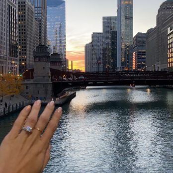 Diamonds - Engagement ring - A hand with an engagement ring is positioned in the foreground, partially obscuring the view of a city skyline with tall modern buildings and a river. The sunset sky displays warm colors, and the scene captures an urban landscape with reflections on the water. - Jewelry Near Me - Chicago, Illinois