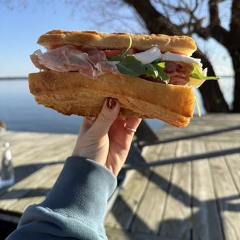 Alimentari - A person holds a sizable sandwich with meats, greens, and bread, outdoors near a lake or river with a tree and a wooden deck in the background. - International Grocery Near Me - Madison, Wisconsin