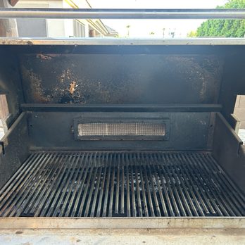 BBQ Cleaning Guru - Before cleaning and in need of serious help. - A used outdoor barbecue grill with its lid open, revealing a rusty interior and grill grates. The grill appears to be in need of cleaning and maintenance, with visible rust and debris inside. It is situated outdoors on a table, with a background of a building and trees. - Grill Services Near Me - Phoenix, Arizona