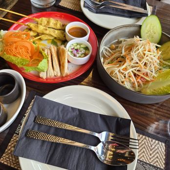 Thai Table - Time to eat! - A Thai restaurant meal featuring chicken satay skewers, small sandwiches, and a large bowl of papaya salad with cucumber and lettuce, served on a wooden table. - Thai Near Me - Frederick, Maryland
