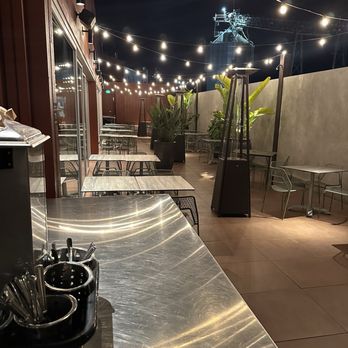 Picante - Redesigned outdoor patio - An outdoor dining area at night with string lights hanging overhead. The patio has several tables and chairs, some with metal frames and others with wooden surfaces. Tall patio heaters are positioned throughout the space, and large potted plants are placed along the concrete wall. The background shows a dark sky and industrial structures, indicating an urban setting. The area appears to be part of a restaurant or bar with a modern, open-air design. - Mexican Near Me - Berkeley, California