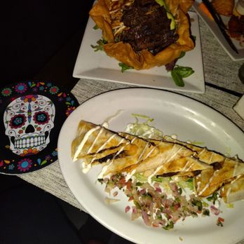 El Patron - The image features two dishes on a table at a restaurant. The first dish is a quesadilla with melted cheese, drizzled with white and dark sauces, served with chopped vegetables and shredded lettuce. The second dish is a Colombian bandeja paisa served in a decorative bowl, containing grilled meat, plantains, and other traditional ingredients. A colorful skull-themed plate is also visible on the table, indicating a festive or cultural setting. - Mexican Near Me - Flushing, New York