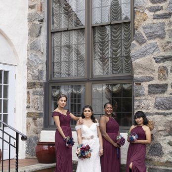 Douglaston Manor - Four women, including a bride in a white gown, stand on steps outside a stone building with large windows and curtains. - Venues & Event Spaces Near Me - Douglaston, New York