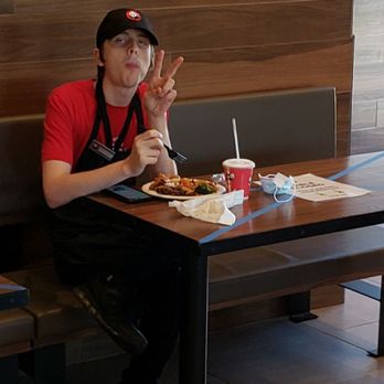 Panda Express - Employee seems to be happy that he can sit and eat but I can't. - A young man in a red shirt and black cap sits at a restaurant table with food and a drink, making a peace sign with his hand. - Chinese Near Me - Kaufman, Texas