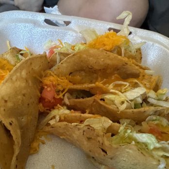 Perico's Albuquerque - Beef tacos - A foam takeout container contains beef tacos with crispy taco shells filled with shredded lettuce, diced tomatoes, and shredded cheese. The tacos are arranged in the container, which is held in front of a person. The background shows part of a person's arm and clothing, indicating a casual dining or takeout setting. - Mexican Near Me - Albuquerque, New Mexico