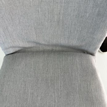 LJC Style Upholstery - Puckering. - The image depicts a gray upholstered chair with a textured fabric surface, showing the seat and backrest. The fabric has a fine, linear pattern. Part of a shoe is visible at the edge of the frame, indicating a person may be nearby. The setting appears to be indoors, possibly in an office or waiting area. - Furniture Reupholstery Near Me - Hanover, Massachusetts