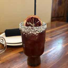 Midnight margarita - A tall glass of dark cocktail with a salted rim and a dried citrus slice garnish, placed on a wooden table in a restaurant setting.