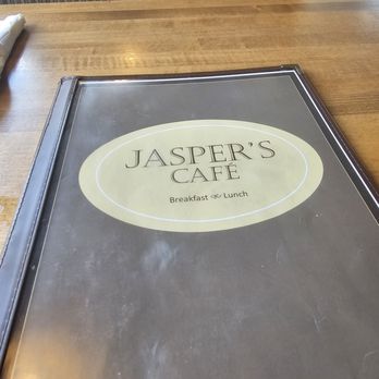 Jasper's Cafe - A menu from Jasper's Café, displaying the restaurant's name and offerings for breakfast and lunch, is positioned on a wooden table. The menu has a black border and a light-colored oval logo with the café's name. The table surface is smooth and wooden, with a patterned fabric visible in the background. - Breakfast & Brunch Near Me - Glenview, Illinois
