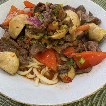 Laghman Express - A plate of stir-fried beef with sliced mushrooms, chopped carrots, and green onions, served over a bed of noodles with a light sauce. The dish is presented on a white plate placed on a green textured placemat, showcasing a colorful and savory Asian-inspired meal. - Halal Near Me - Brooklyn, New York