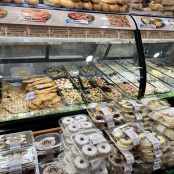 Bagel Boy - The image shows a glass display case filled with salads, fried foods, and creamy dishes, along with packaged baked goods and cookies arranged on the counter in front. The case is part of a deli or bakery offering a variety of prepared foods and desserts. - Bagels Near Me - Brooklyn, New York