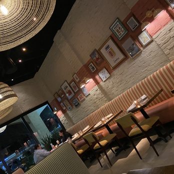 NiMo - Dining room - The image shows the interior of a restaurant with a long, striped upholstered banquette along a white brick wall decorated with framed artwork. Small tables with chairs are arranged in front of the banquette, set with napkins and glasses. Large woven pendant lights hang from the ceiling, providing warm lighting. A glass window or door is visible at the far end, with a couple of patrons seated and engaged in conversation. The decor includes a mix of modern and cozy elements, creating a casual dining atmosphere. - Salad Near Me - Tequesta, Florida