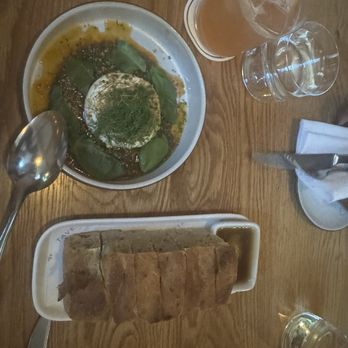 Tavernetta - Barrata and focaccia bread - The image shows a wooden table with a plate of stuffed green peppers topped with herbs and sauce, a sliced loaf of bread with dipping sauce, and several drinks including a cocktail and glasses of water. Cutlery and napkins are also visible, indicating a dining setting at a restaurant. - Italian Near Me - Denver, Colorado