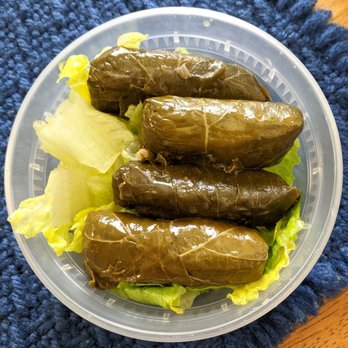Caravan Grill - Stuffed Grape Leaves - Four stuffed grape leaves are placed on a bed of light green lettuce inside a round, transparent plastic container. The grape leaves are dark green and glossy, indicating they are preserved or cooked. The container rests on a textured blue surface, possibly a cloth or mat, with part of a wooden surface visible in the top right corner. - Halal Near Me - Tucson, Arizona
