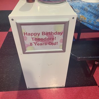 Flying Squirrel - My son, Theo, had his own table and set-up - A white stand displays a birthday message for Theodore's 8th birthday, with party decorations and tables in the background. - Trampoline Parks Near Me - Ottawa, Ontario