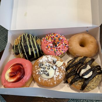 Donerds Donuts - Dubai chocolate  Homer Original Raspberry cheesecake Cannoli Smores - A box contains six donuts with different toppings, including chocolate, sprinkles, pink glaze, and powdered sugar, on a wooden table. - Donuts Near Me - Bethlehem, Pennsylvania