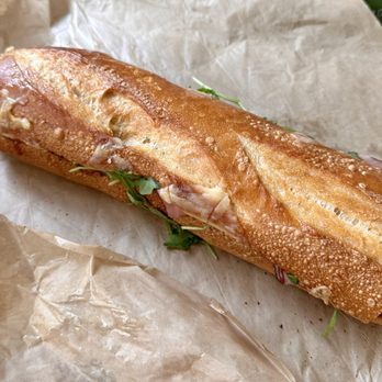 Panera Bread - Ham & Gouda Baguette - A baguette sandwich with a crispy, golden-brown crust contains slices of ham, gouda cheese, and fresh greens. The sandwich is resting on crumpled parchment paper, highlighting its fresh and artisanal appearance. - Sandwiches Near Me - Needham, Massachusetts