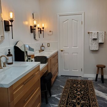 Kobolt Construction - a bathroom with two sinks and a rug - A bathroom features two white vanities with brass fixtures, wall-mounted light fixtures, and a mirror. Towels hang on a rack, and a small stool is present. - General Contractors Near Me - Fullerton, California
