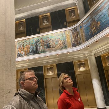Inside Chicago Walking Tours - Two visitors, a man and a woman, are looking upward inside a building featuring a decorative mosaic mural on the upper level. The interior includes gold-framed wall niches with sculptures and black marble walls with gold accents. The mosaic depicts a historical or mythological scene, and the setting appears to be a museum, gallery, or historic building with classical architectural elements. - Architectural Tours Near Me - Chicago, Illinois