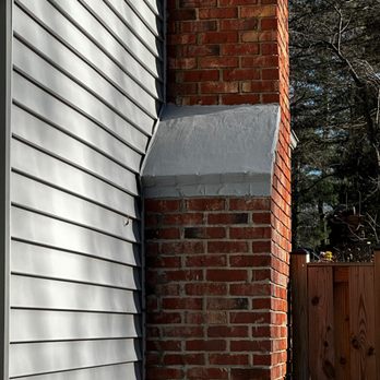 Chimpro chimney sweeps - After - The image shows a close-up of a brick chimney with gray metal flashing installed at its base, attached to a house with white vinyl siding. The background includes trees and a wooden fence, indicating an outdoor residential setting. The lighting suggests it is daytime with sunlight illuminating the scene. - Chimney Sweeps Near Me - Alexandria, Virginia