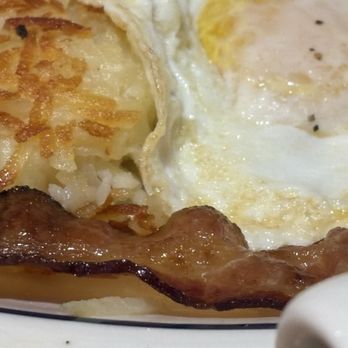 IHOP - The image shows a breakfast plate with crispy hash browns, a fried egg topped with black pepper, and several strips of cooked bacon. The hash browns are golden and shredded, the egg has a runny yolk, and the bacon appears crispy and browned. The food is served on a white plate with a black rim, and the focus is on the food's textures and colors. - Breakfast & Brunch Near Me - Union, New Jersey