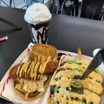 Level Zero - A food tray contains two sandwiches topped with sliced jalapeños and a creamy sauce, a chocolate milkshake with whipped cream in a branded cup, and a cookie. The tray is placed on a black table in a casual dining setting with chairs visible in the background. - Halal Near Me - Dearborn, Michigan