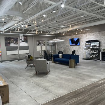 MSI - the inside of a furniture store - The image shows an interior showroom with a high ceiling and exposed ductwork. There is a seating area with a blue sofa, a gray armchair, and a small side table. The wall displays various sample materials and finishes, and a television is mounted on a brick-textured wall. The space is illuminated by string lights and ceiling spotlights, with a vending machine and storage cabinets visible in the background. - Building Supplies Near Me - Hatboro, Pennsylvania