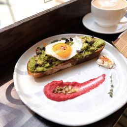 Avo Toast - A white plate holds a slice of toasted bread topped with mashed avocado, microgreens, and a fried egg with a runny yolk. There is a small dollop of cream or sauce and a sprinkle of seeds on the plate. A red sauce smear and a small herb sprig are also present on the plate. In the background, a white cup filled with a frothy coffee beverage is visible. The setting appears to be near a window with natural light, on a wooden surface.