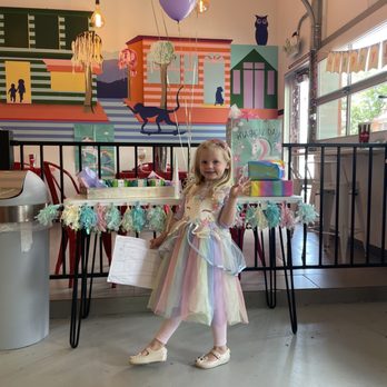 Scoops - a little girl in a dress standing in front of a table with balloons - A girl in a colorful, pastel rainbow dress stands in front of a decorated table at a birthday celebration, holding a peace sign. - Ice Cream & Frozen Yogurt Near Me - Denver, Colorado
