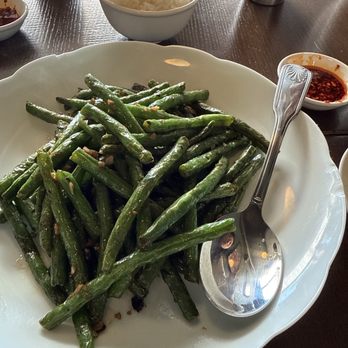 Dumpling Wei - String beans with rice - A white, scalloped-edge plate contains stir-fried green beans with visible garlic pieces, placed on a dark wooden table. Surrounding the plate are a bowl of white rice, small bowls of sauce, and metal cups, indicating a meal setting. The image captures a casual dining scene with Asian cuisine elements. - Chinese Near Me - Pasadena, California