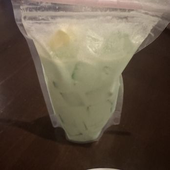Decades - Shamrock in a shake pouch drink - A clear plastic bag contains a greenish-colored drink with ice cubes and lemon slices inside. A pink straw is inserted into the bag, which is resting on a dark wooden surface. Part of a white plate with some food is visible at the bottom of the image. - Arcades Near Me - Lancaster, Pennsylvania