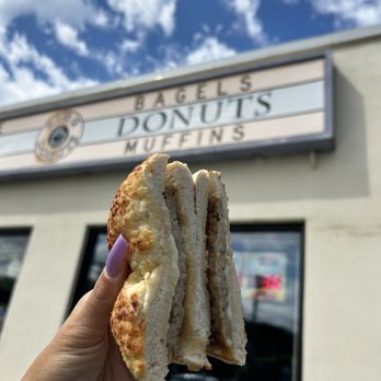 King Donuts - a hand holding a half eaten sandwich - A person holds a sandwich with bread and filling outside a donut shop, with a sign reading 'Bagels, Donuts, Muffins' in the background and a partly cloudy sky. - Donuts Near Me - Newington, Connecticut