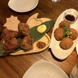 Karaage and croquettes - The image shows two plates of fried food on a wooden table. One plate contains fried chicken pieces with a dollop of mayonnaise and a small pile of seasoning or spice mix. The other plate has three croquettes garnished with a small green leaf and yellow flowers, with a small bowl of dipping sauce. There is also a glass of water and an empty white plate visible in the scene.