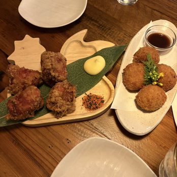 Tsuruya - Karaage and croquettes - The image shows two plates of fried food on a wooden table. One plate contains fried chicken pieces with a dollop of mayonnaise and a small pile of seasoning or spice mix. The other plate has three croquettes garnished with a small green leaf and yellow flowers, with a small bowl of dipping sauce. There is also a glass of water and an empty white plate visible in the scene. - Ramen Near Me - Berkeley, California