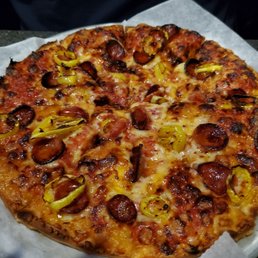 Limguica and hot pepper pizza.  1/2 price on Mondays - A freshly baked pizza featuring slices of sausage, yellow peppers, and melted cheese, placed on a white paper liner. The pizza has a golden-brown crust and appears to be served in a casual dining setting, possibly at a coastal restaurant or sports bar known for seafood and pizza offerings.
