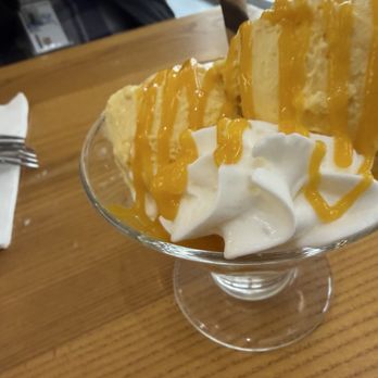 Mango Mango Dessert - The image shows a dessert in a glass dish containing whipped cream topped with caramel sauce. Slices of cake are placed on top, with a chocolate wafer stick inserted into the cake. The dessert is on a wooden table near a window, with a napkin and fork visible nearby. The setting appears to be a casual dining or dessert establishment. - Desserts Near Me - Plainview, New York