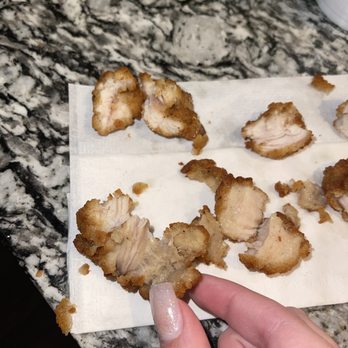 Chick-fil-A - ows a person holding a piece of food - Several pieces of fried chicken are arranged on a paper towel, which is on a granite countertop. The chicken pieces vary in size and shape, with a golden-brown crispy coating. A person's hand with glittery nail polish is holding one piece of chicken. The background includes a white cup and a small yellow object on the countertop. - Fast Food Near Me - Kyle, Texas