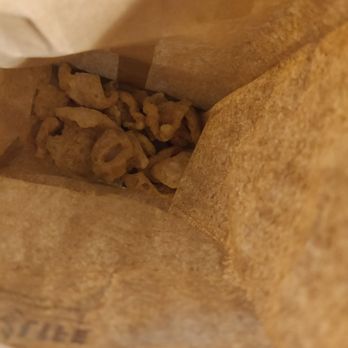 Thai Lada - Pork Rinds with Boat Noodles - The image shows the interior of a brown paper bag with pork rinds inside. The pork rinds are light brown, irregularly shaped, and appear crispy. The bag is partially open, revealing the snack inside, and the photo is taken from above, focusing on the contents. - Thai Near Me - Austin, Texas